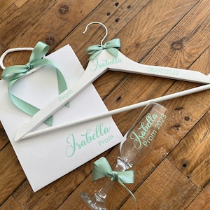 May include: White gift bag with a green ribbon and the word "Prom" written on it. A white wooden hanger with the name "Isabella" and the date "08/07/2023" written on it. A clear glass with the name "Isabella" and the words "Prom 2023" written on it.