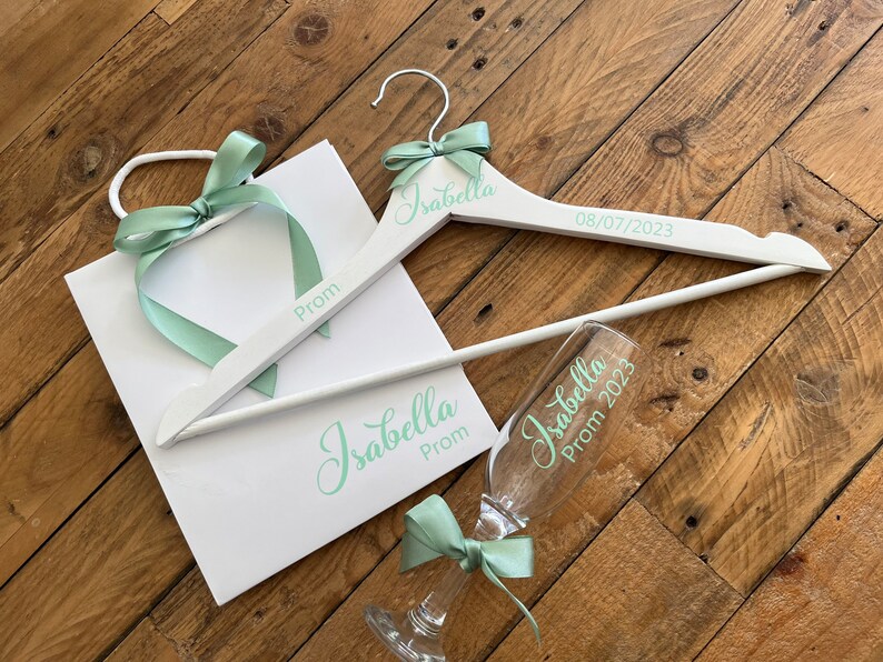 May include: White gift bag with a green ribbon and the word "Prom" written on it. A white wooden hanger with the name "Isabella" and the date "08/07/2023" written on it. A clear glass with the name "Isabella" and the words "Prom 2023" written on it.