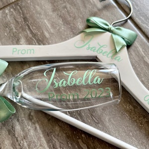 May include: White wooden hanger with a green bow and the text "Isabella Prom 2023" and "08/07/2023". A champagne flute with a green bow and the text "Isabella Prom 2023" is in front of the hanger.