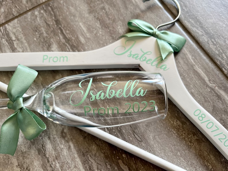 May include: White wooden hanger with a green bow and the text "Isabella Prom 2023" and "08/07/2023". A champagne flute with a green bow and the text "Isabella Prom 2023" is in front of the hanger.