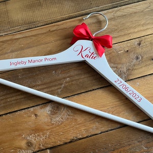 May include: White wooden hanger with a red bow and the text "Katie" and "Ingleby Manor Prom 27/06/2023" in red.