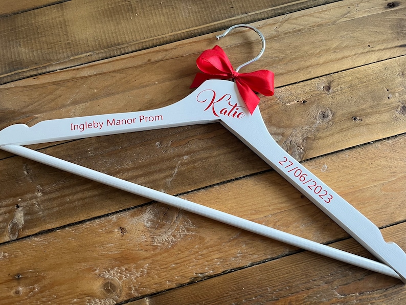 May include: White wooden hanger with a red bow and the text "Katie" and "Ingleby Manor Prom 27/06/2023" in red.