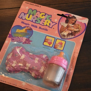 May include: A pink and white Magic Nursery Magic Milk Bottle toy packaging with a picture of a doll and a clear bottle with a pink lid. The packaging says "Magic Nursery Magic Milk Bottle" and "For Ages Over 3".