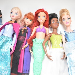 May include: A collection of seven dolls, each representing a different character. The dolls are dressed in colorful outfits, including gowns, dresses, and tops. The dolls have various skin tones and hairstyles, and are arranged in a row.