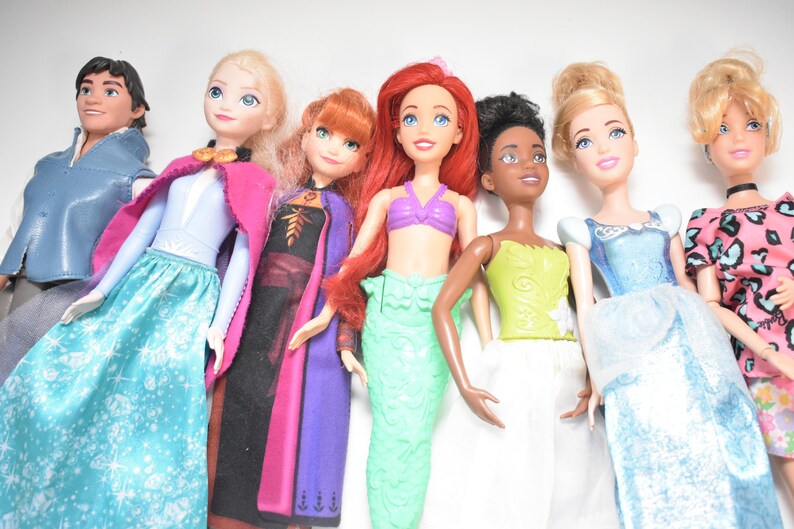 May include: A collection of seven dolls, each representing a different character. The dolls are dressed in colorful outfits, including gowns, dresses, and tops. The dolls have various skin tones and hairstyles, and are arranged in a row.