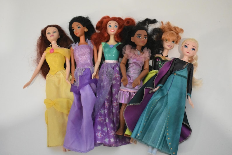 May include: A collection of seven dolls, each dressed in colorful gowns. The dolls have various hair colors, including red, black, and blonde. The dresses are in shades of yellow, purple, green, and blue.