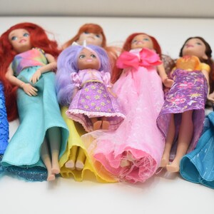 May include: A collection of seven dolls arranged in a row. The dolls feature various hair colors including red, purple, and brown, and wear diverse outfits such as blue mermaid tails, pink gowns, and patterned dresses in shades of yellow and purple.