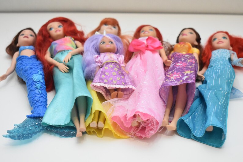 May include: A collection of seven dolls arranged in a row. The dolls feature various hair colors including red, purple, and brown, and wear diverse outfits such as blue mermaid tails, pink gowns, and patterned dresses in shades of yellow and purple.