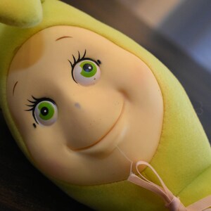 May include: A plush green and yellow caterpillar toy with a smiling face and large green eyes. The toy has a pink ribbon tied around its neck.