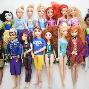 May include: A collection of fashion dolls featuring various characters with detailed outfits and hairstyles. The dolls include characters with blonde, red, and black hair, wearing dresses, tops, and pants in various colors. The dolls are standing on a white surface.