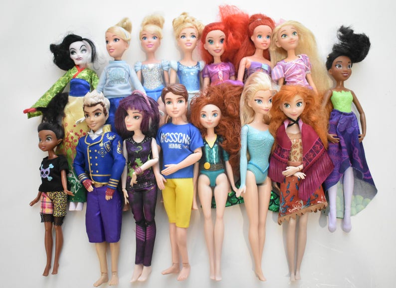 May include: A collection of fashion dolls featuring various characters with detailed outfits and hairstyles. The dolls include characters with blonde, red, and black hair, wearing dresses, tops, and pants in various colors. The dolls are standing on a white surface.