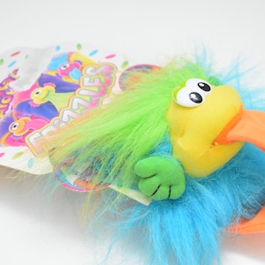 May include: A colorful plush toy parrot with a yellow beak and a blue body. The parrot has green and blue feathery hair and large white eyes. The toy is attached to a cardboard backing with the text "Arcade Frazzles".