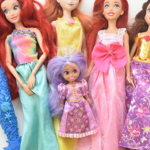 May include: A collection of five dolls featuring various outfits and hair colors. One doll has long red hair and a blue mermaid tail, another has orange hair in a yellow dress, one has red hair in a pink gown, one has brown hair, and a small doll has purple hair.
