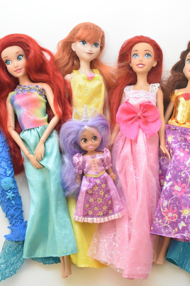 May include: A collection of five dolls featuring various outfits and hair colors. One doll has long red hair and a blue mermaid tail, another has orange hair in a yellow dress, one has red hair in a pink gown, one has brown hair, and a small doll has purple hair.