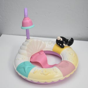 May include: A colorful, pastel-colored inflatable ring toy with a seashell design. It features a pink whale-shaped seat, a small black and white whale figurine, and a pink and purple sunshade. The ring is decorated with a starfish and other sea-themed details.