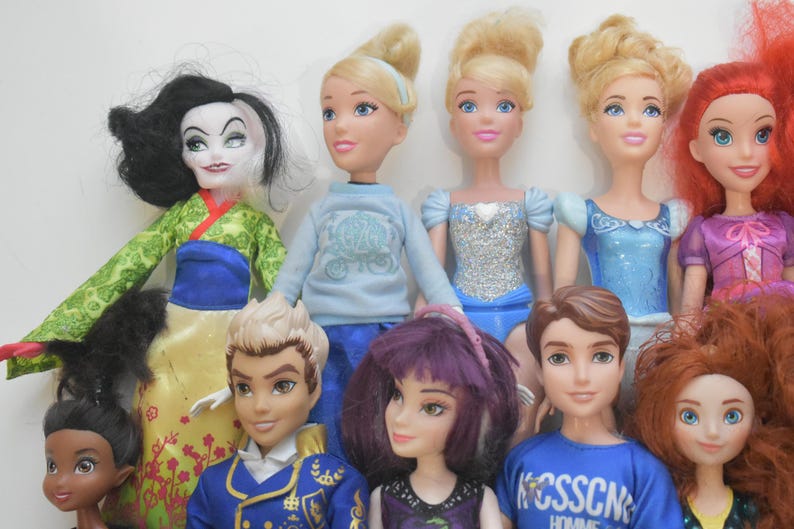 May include: A collection of dolls featuring various characters. The dolls have different hair colors and styles, and are wearing a variety of outfits. The dolls are arranged in a group, with some standing in the front and others in the back.