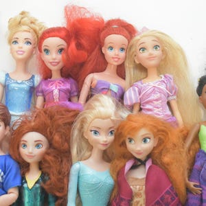 May include: A collection of dolls featuring various characters with detailed facial features and colorful outfits. The dolls have long hair in shades of blonde, red, and black. Some dolls are wearing dresses in blue, pink, and purple.