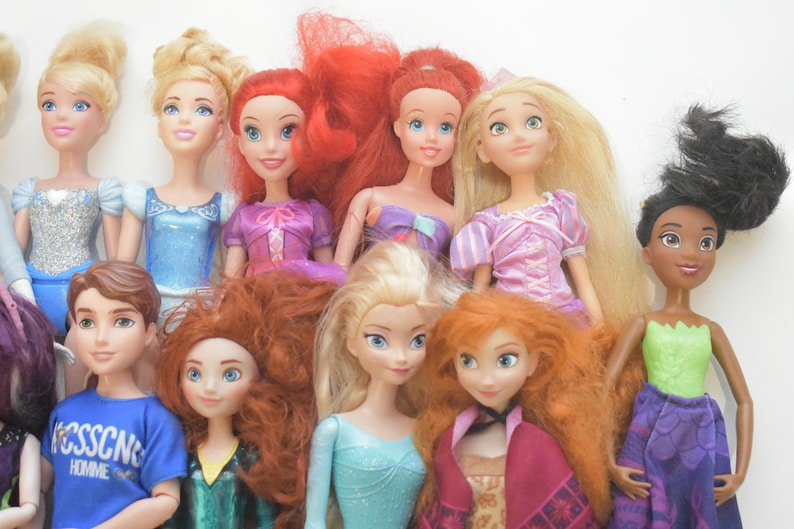 May include: A collection of dolls featuring various characters with detailed facial features and colorful outfits. The dolls have long hair in shades of blonde, red, and black. Some dolls are wearing dresses in blue, pink, and purple.