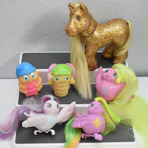 May include: A collection of vintage My Little Pony toys, including a gold glitter pony, a pink bird with a teacup, a purple bird with a crown, a green caterpillar, and a blue and pink caterpillar. The toys are arranged on a white tiered shelf.