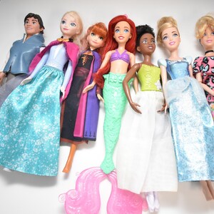 May include: A collection of seven dolls, including a male doll in a blue jacket and gray pants, and several female dolls in various gowns and dresses. The dolls have different skin tones and hair colors, with some wearing tiaras.
