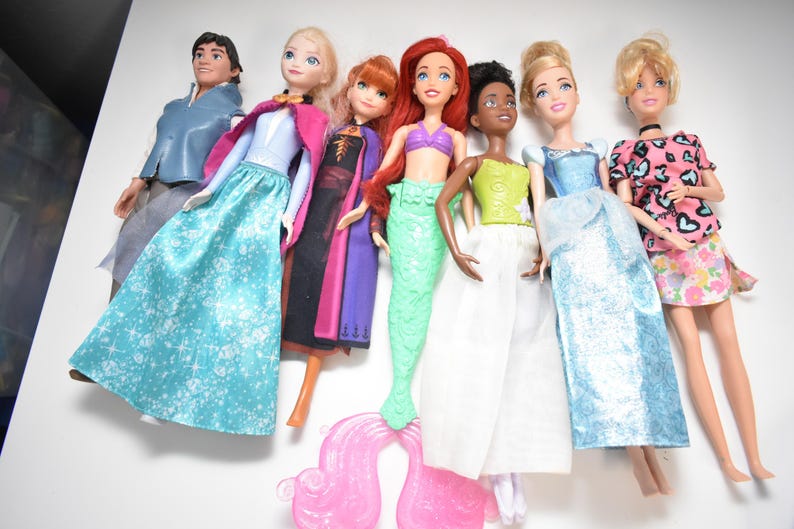 May include: A collection of seven dolls, including a male doll in a blue jacket and gray pants, and several female dolls in various gowns and dresses. The dolls have different skin tones and hair colors, with some wearing tiaras.