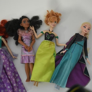 May include: Several dolls are arranged on a white surface. The dolls have various skin tones and hair colors, including red, black, and blonde. They are wearing colorful dresses in shades of purple, green, and blue. The dolls appear to be based on animated characters.