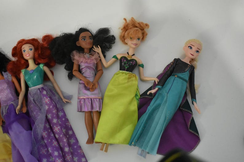 May include: Several dolls are arranged on a white surface. The dolls have various skin tones and hair colors, including red, black, and blonde. They are wearing colorful dresses in shades of purple, green, and blue. The dolls appear to be based on animated characters.