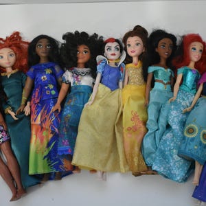 May include: A collection of ten dolls, each dressed in colorful gowns and outfits. The dolls have various skin tones and hairstyles, including red, black, and blonde hair. The dolls are arranged in a row, showcasing their diverse designs.