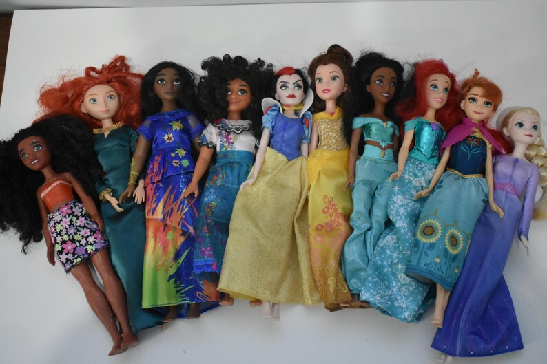 May include: A collection of ten dolls, each dressed in colorful gowns and outfits. The dolls have various skin tones and hairstyles, including red, black, and blonde hair. The dolls are arranged in a row, showcasing their diverse designs.