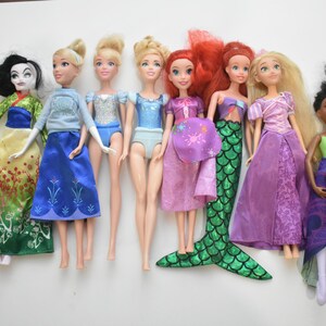 May include: A collection of eight dolls, each dressed in colorful outfits. The dolls include characters with long hair, dresses, and unique accessories. The dolls are arranged in a row against a white background.