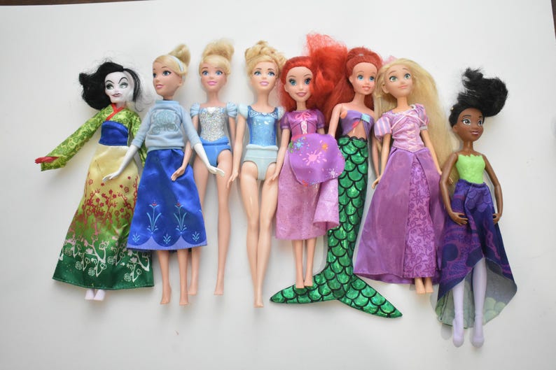 May include: A collection of eight dolls, each dressed in colorful outfits. The dolls include characters with long hair, dresses, and unique accessories. The dolls are arranged in a row against a white background.
