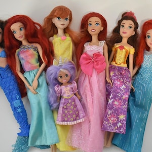 May include: A collection of six dolls featuring various outfits and hair colors. The dolls include figures with red, brown, and purple hair, wearing dresses in blue, yellow, pink, and purple, with one doll featuring a mermaid tail and another a unicorn horn.