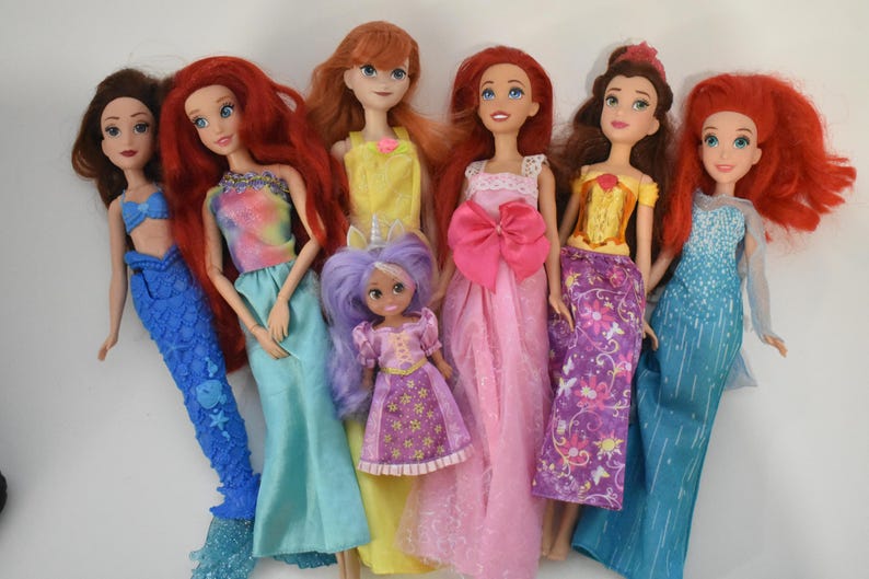 May include: A collection of six dolls featuring various outfits and hair colors. The dolls include figures with red, brown, and purple hair, wearing dresses in blue, yellow, pink, and purple, with one doll featuring a mermaid tail and another a unicorn horn.