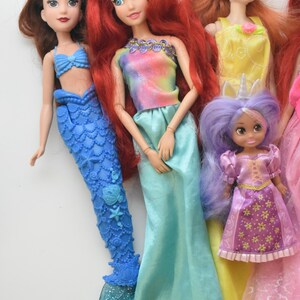 May include: A collection of five dolls featuring various outfits and hair colors. One doll has a blue mermaid tail, another wears a tie-dye top with a turquoise skirt, one has a yellow dress, a small doll has purple hair, and one wears a pink dress.