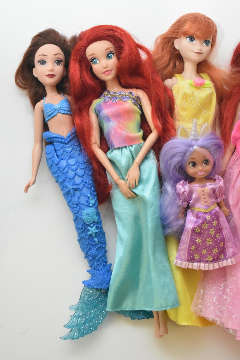 May include: A collection of five dolls featuring various outfits and hair colors. One doll has a blue mermaid tail, another wears a tie-dye top with a turquoise skirt, one has a yellow dress, a small doll has purple hair, and one wears a pink dress.