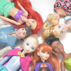 May include: A collection of Disney princess dolls, including Ariel, Elsa, Anna, and others, arranged in a circular pattern. The dolls feature vibrant colors and detailed outfits, showcasing their iconic looks. The dolls are lying on a white surface.