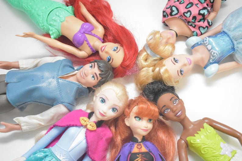 May include: A collection of Disney princess dolls, including Ariel, Elsa, Anna, and others, arranged in a circular pattern. The dolls feature vibrant colors and detailed outfits, showcasing their iconic looks. The dolls are lying on a white surface.