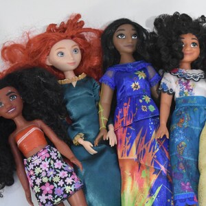 May include: A collection of dolls featuring various characters with distinct hairstyles and colorful outfits. The dolls include a character with red hair in a teal dress, and another with black hair in a blue dress with floral accents.