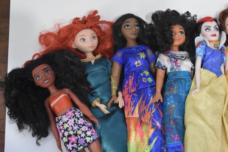 May include: A collection of dolls featuring various characters with distinct hairstyles and colorful outfits. The dolls include a character with red hair in a teal dress, and another with black hair in a blue dress with floral accents.