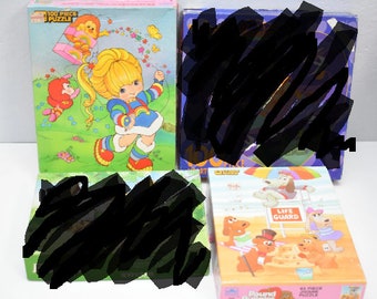 Puede incluir: Cuatro cajas de rompecabezas vintage. Una caja presenta una niña de dibujos animados con un tema de arcoíris, el texto "100 Piece Puzzle". Otra caja tiene un perro salvavidas de dibujos animados. Las otras dos cajas están parcialmente oscurecidas.