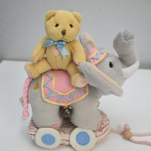 May include: A plush toy featuring a yellow teddy bear riding a gray elephant. The elephant has a pink and blue saddle, a decorative hat, and a small bell. The toy is on a wheeled base with a pink pull string.