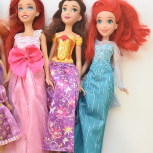 May include: Three dolls with distinct features. The first has long red hair and a pink gown with a large bow. The second has brown hair, a yellow bodice, and a purple floral skirt. The third has red hair and a blue gown with white vertical lines.