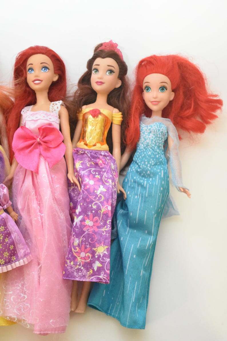 May include: Three dolls with distinct features. The first has long red hair and a pink gown with a large bow. The second has brown hair, a yellow bodice, and a purple floral skirt. The third has red hair and a blue gown with white vertical lines.