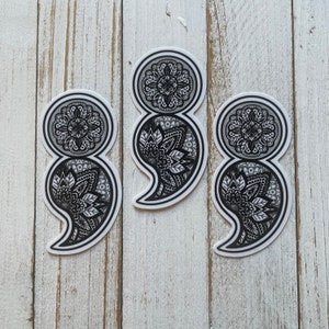 May include: Three black and white semicolon stickers with a mandala design. The stickers are on a white wooden background.