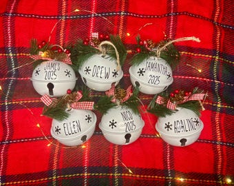 May include: White bell-shaped Christmas ornaments with names and the year 2025. Each ornament is decorated with pine needles, red berries, and a red and white checkered ribbon. The ornaments are on a red and black plaid background.