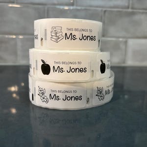 May include: Three white labels with black text and illustrations. The labels read "This belongs to Ms. Jones". The illustrations are a stack of books, an apple, and a flower.