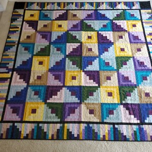 May include: A quilt with a log cabin pattern in shades of purple, blue, green, yellow, and brown. The quilt is made up of squares with a series of triangles in each square.
