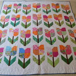May include: A white quilt with a patchwork design of colorful tulips. The tulips are made of various fabrics with floral patterns and geometric shapes. The quilt is quilted with a simple stitch pattern.
