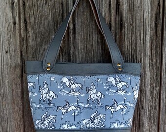 May include: A gray tote bag with a black leather bottom and a blue and white fabric top. The fabric features a repeating pattern of horses and riders jumping over fences.