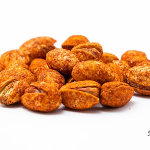 May include: A close-up of a pile of spicy pistachios. The pistachios are roasted and coated in a red chili powder seasoning.